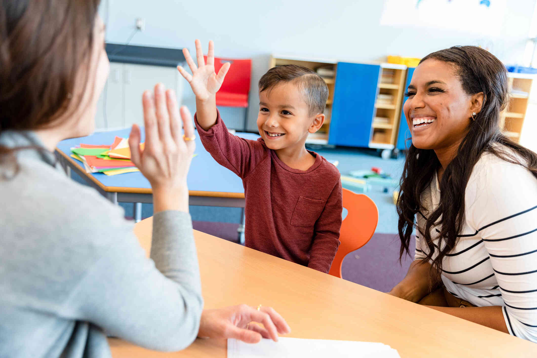 A young boy smiles as he high fives a woman sitting across from him at a classroom desk. An adult woman with long dark hair sits beside the boy and laughs.